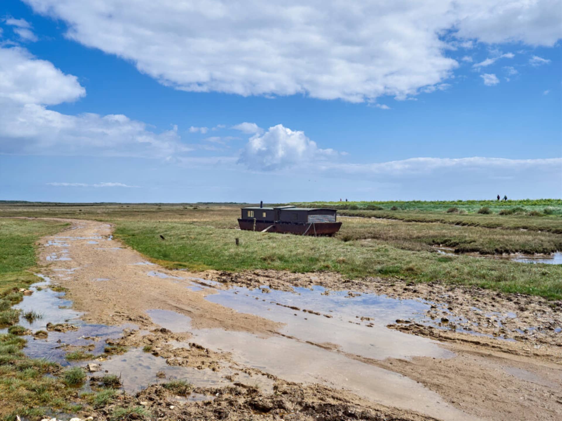 A house boat on Brancaster Marsh with a path and grass in the foreground and people walking on the horizon on a sunny day in Brancaster, Norfolk