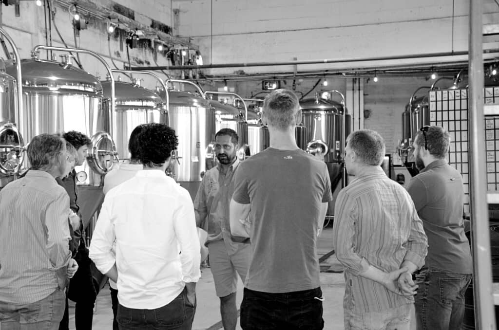 Black and white photo with a tour group with back to camera facing the tour guide in a brewery