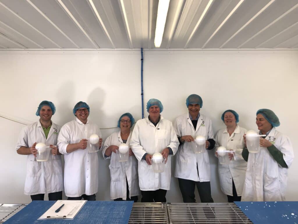 Seven people in white coats and blue hairnets holding plastic jugs with a ball of cheese