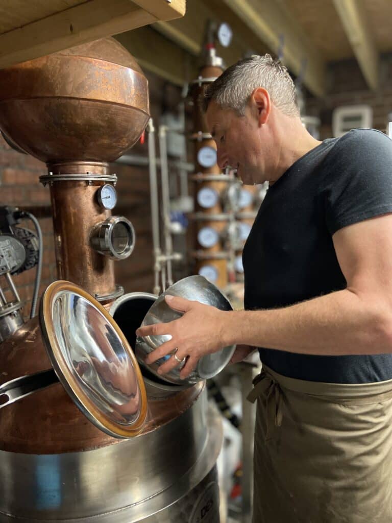 A man pouring from a silver bowl into a distillery barrel.