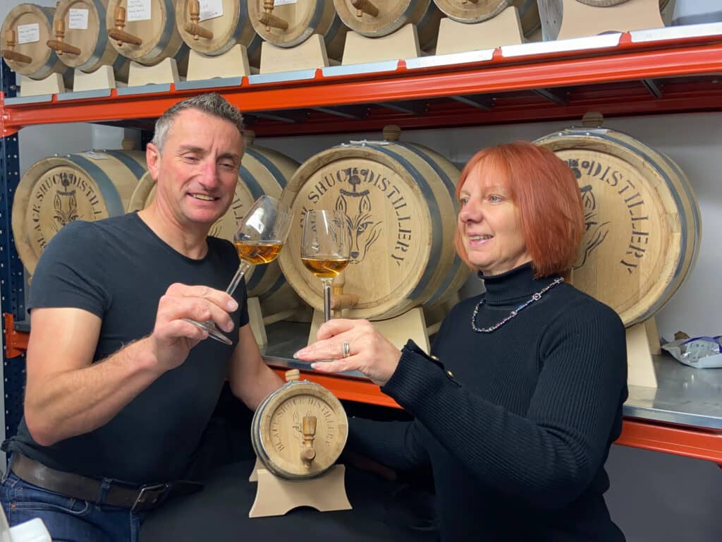 A man and a woman cheering glasses of rum in front of distillery barrels.