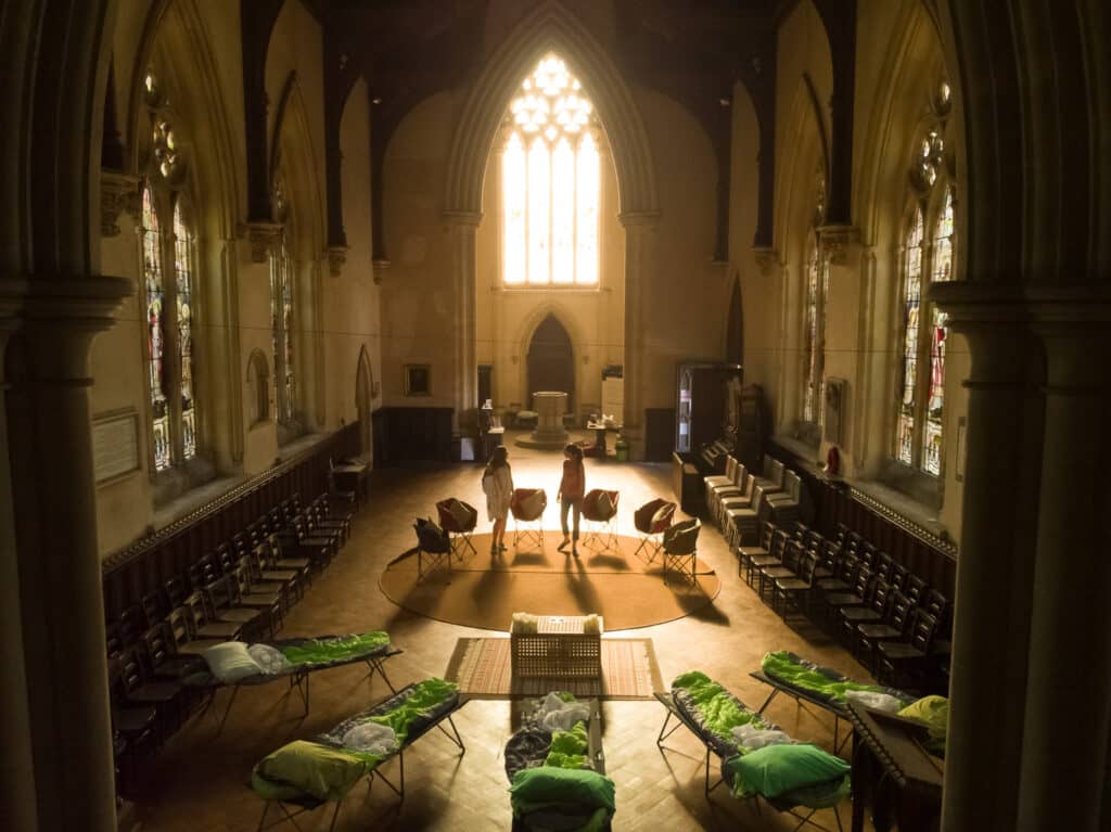 Five camping beds in a semi-circle in a church with sun shining through glass window in background