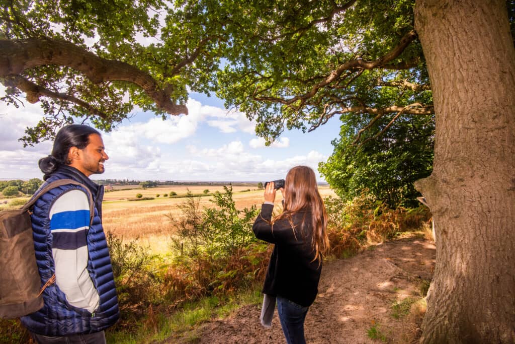 A man and a woman use binoculars to look over some fields on a wooded path at Ken Hill in Norfolk.