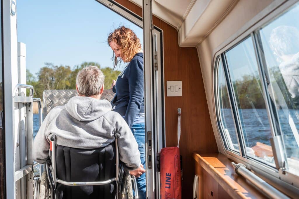 A woman and man in a wheelchairtalk on the decking of a motorboat on the Norfolk Broads.
