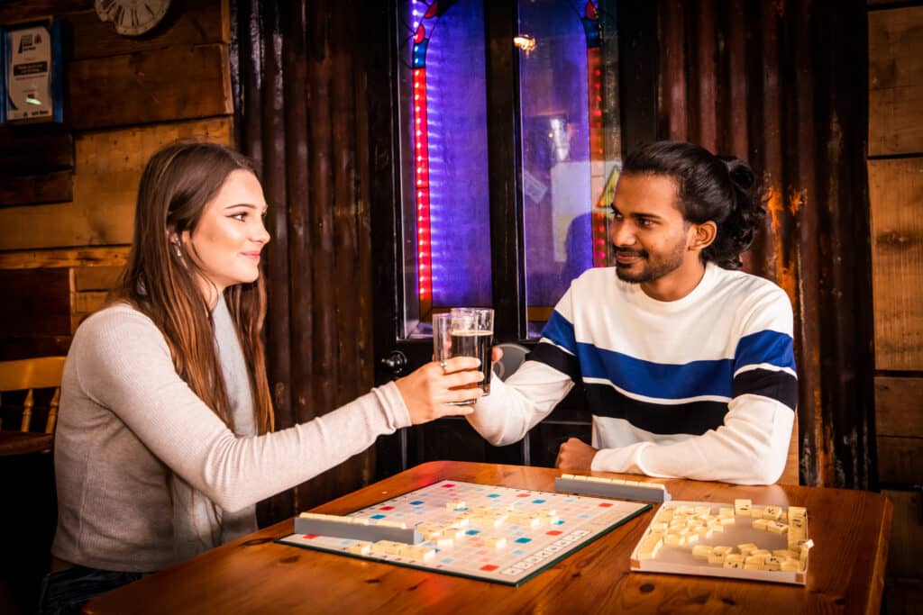 A man and a woman cheers drinks at a wooden table over a game of scrabble in the Nip and Growler Ale House in Norfolk.