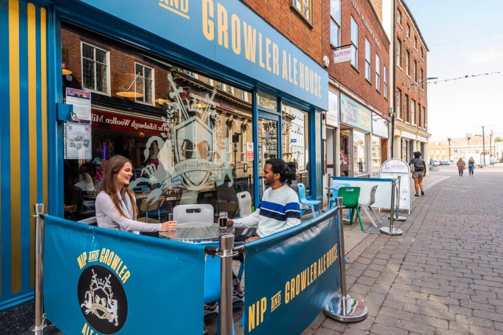 A man and a woman sit at a table outside the Nip and Growler Ale House on a quiet street in Norfolk.