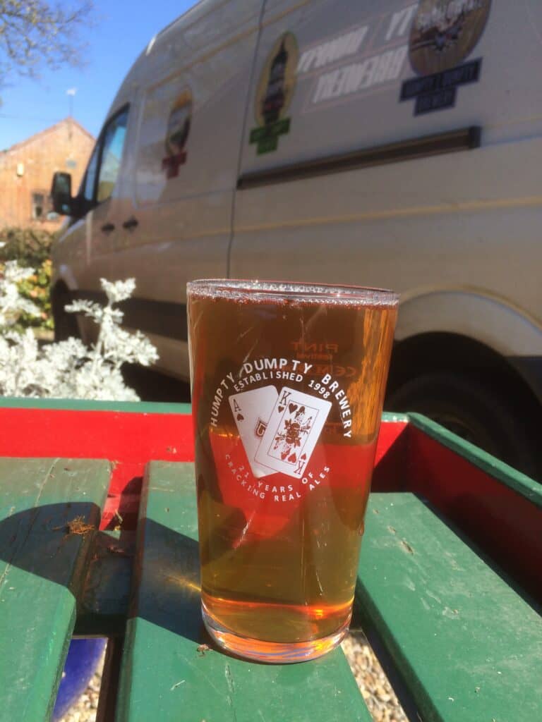 A pint placed on a green bench from Humpty Dumpty Brewery in Norfolk with a van in the background.