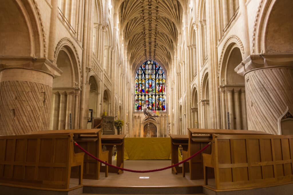 The inside of Norwich Cathedral wirh pews in the foreground and stained glass window in the background.