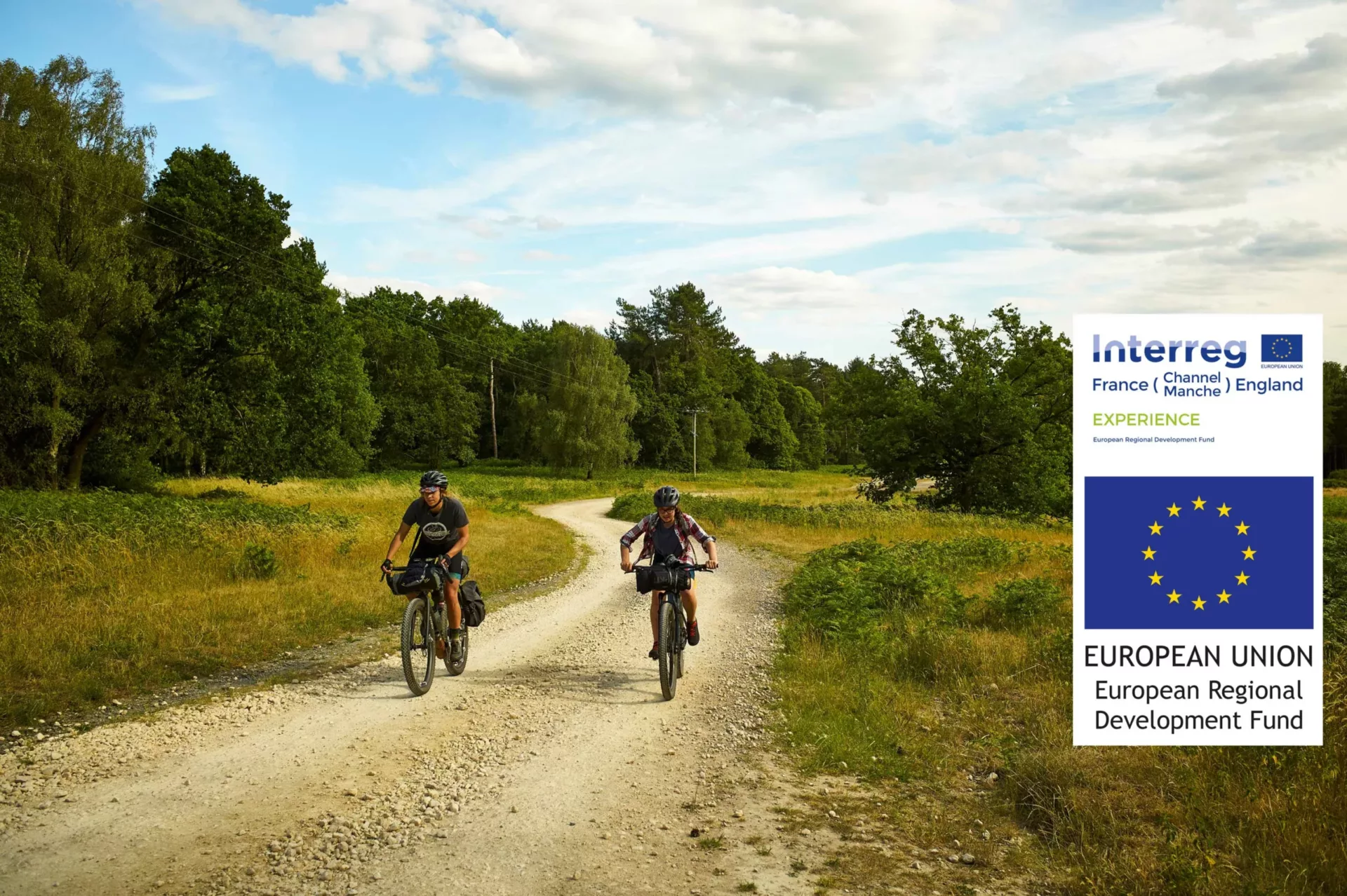 Two people cycle down a rural path with fields on both sides and trees in the background.