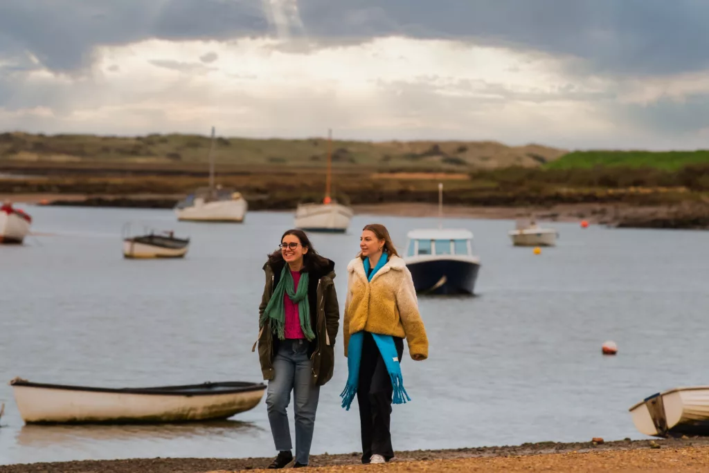 2 young women walking on the beach with water and boats in the background