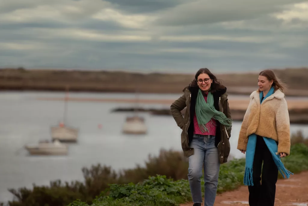 2 young women walking next to the water and few boats in the background