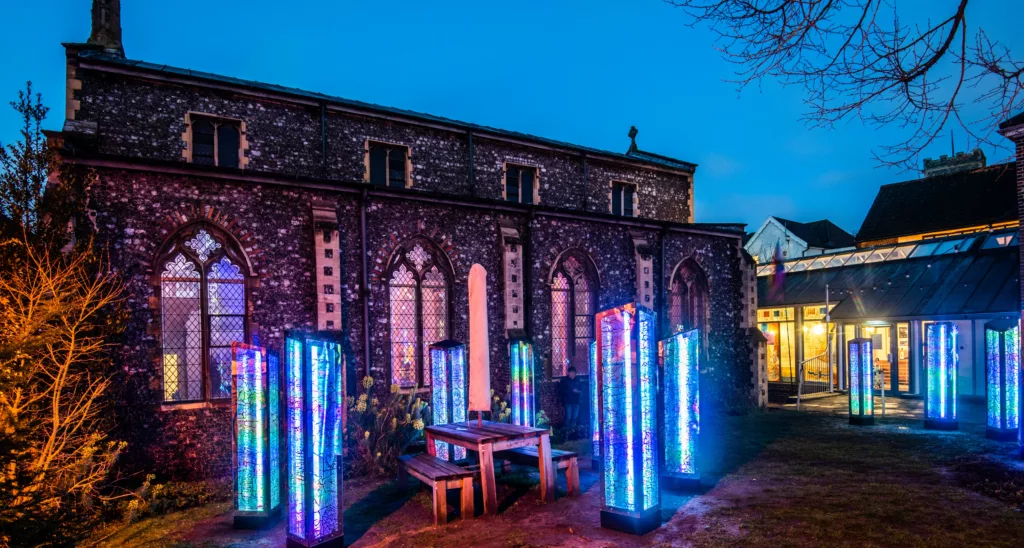 Six art totems lit up blue around a bench outside Norwich Arts Centre