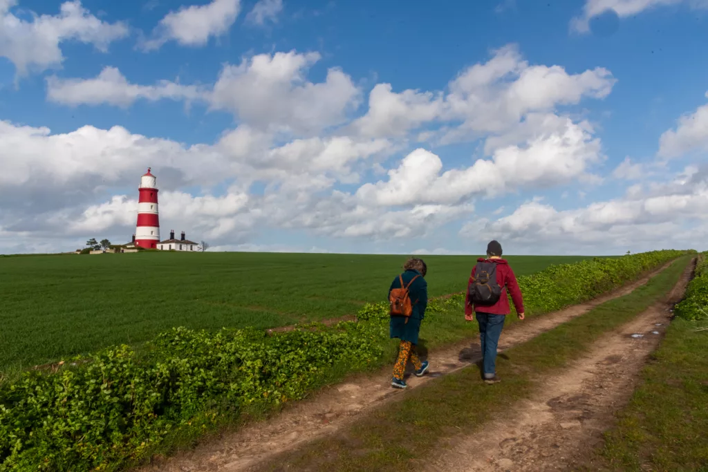 Two people walking on a gravel footpath through fields with Happisburgh Lighthouse in the distance on a sunny day