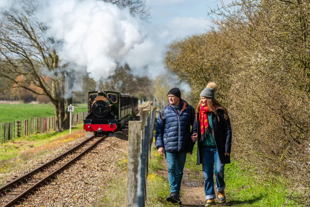 A man and a woman walking on the right as a steam train passes on the left