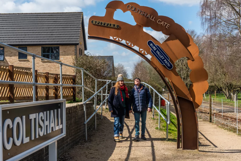 A man and a woman walking under an arch on the Bure Valley Path that reads "Coltishall Gate"