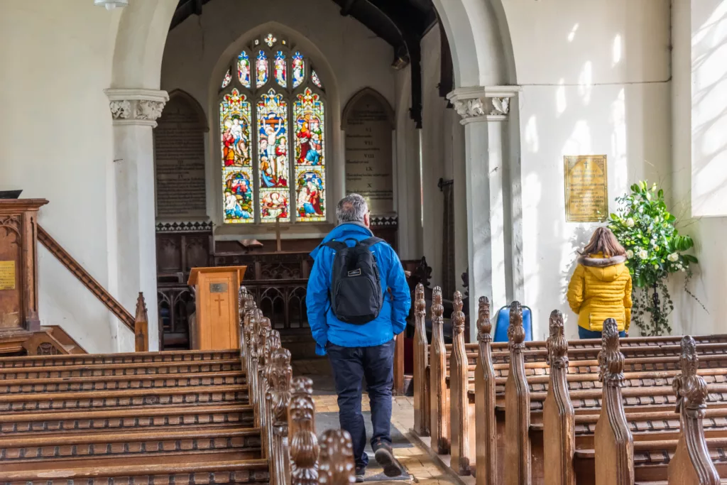 A man and a woman inside All Saints Church, Ashwicken with wooden pews and a stained glass window behind the altar