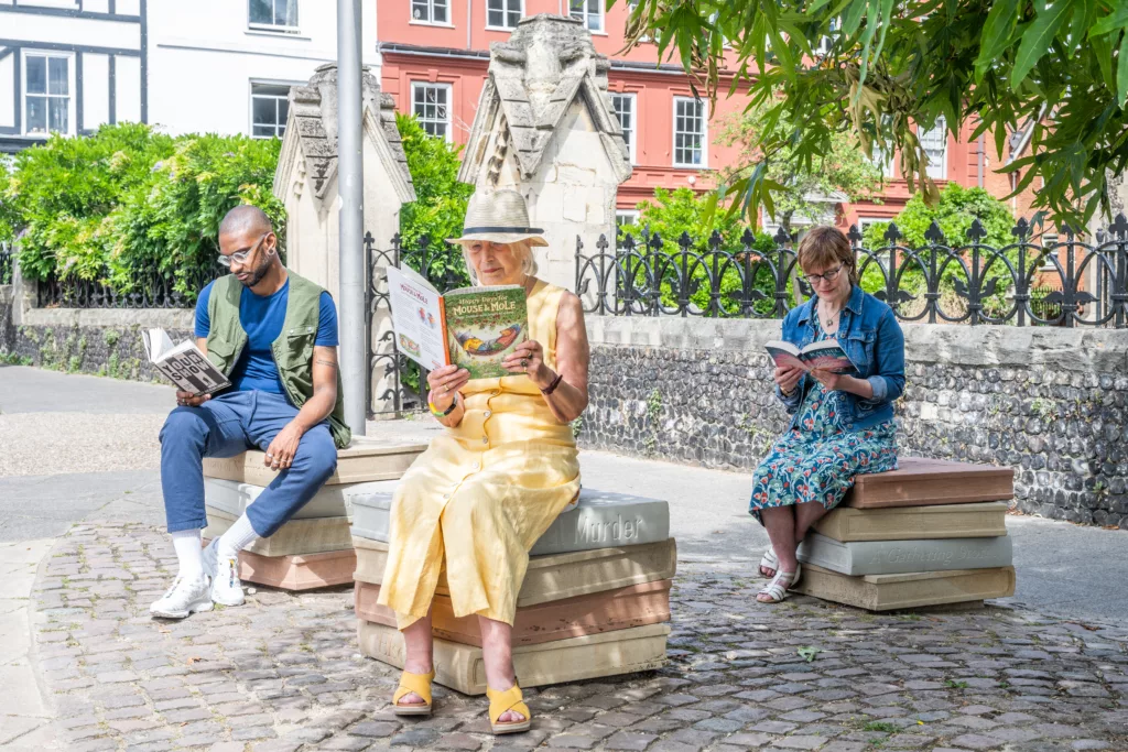 Three people sitting on seats resembling a stack of four books outside St Giles-on-the-Hill, Norwich