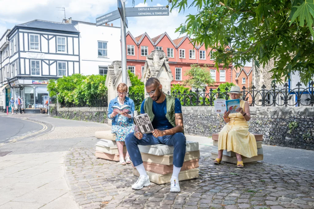 Three people sitting on seats resembling a stack of four books outside St Giles-on-the-Hill, Norwich