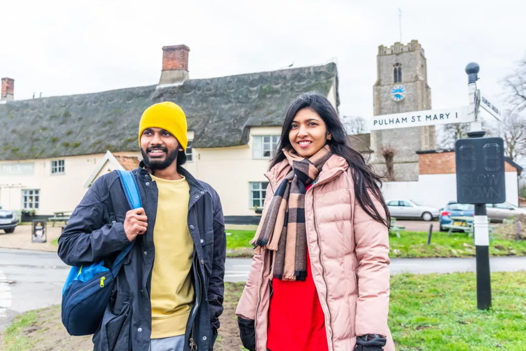 A man and a woman standing in front of a signpost to Pulham St Mary and St Mary Magdelene church in the background